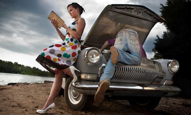 Eine Frau liest ein Buch während ein Mann versucht, das Auto zu reparieren